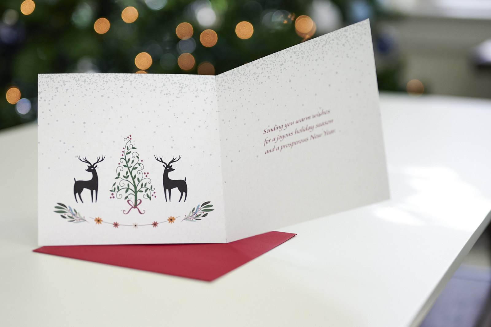 Opened holiday card sitting on top of a red envelope on a table with a blurred Christmas tree in the background.
