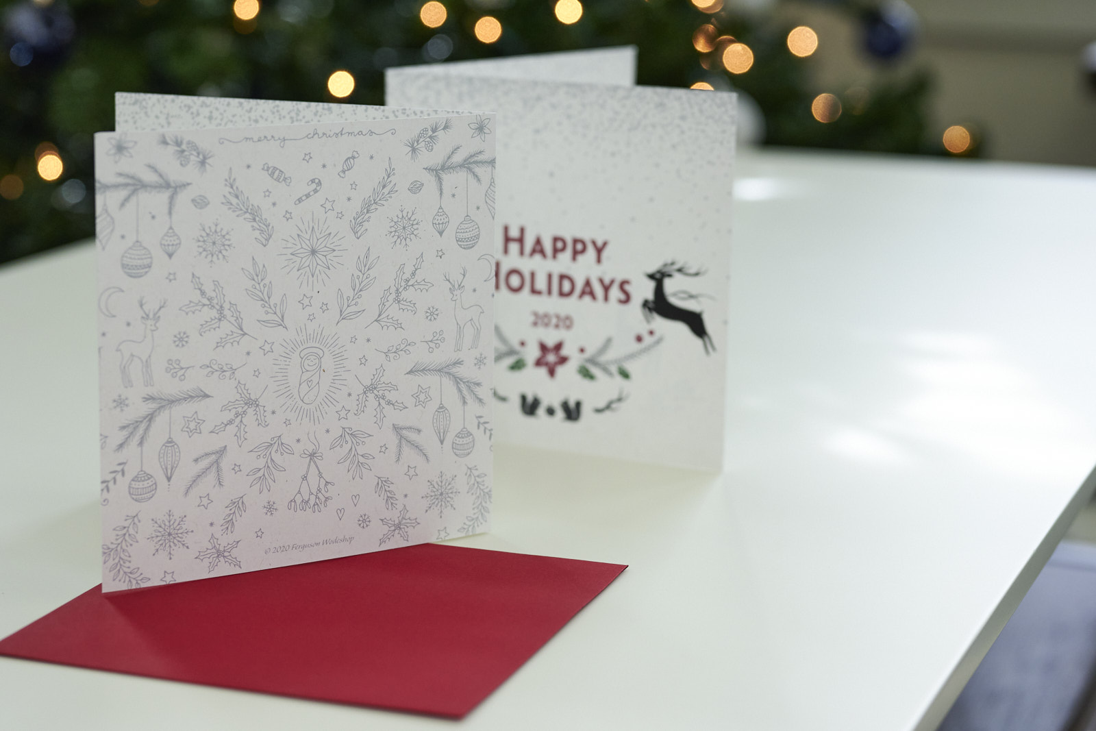 Two holiday cards showing the front and rear views sitting on top of a red envelope on a table with a blurred Christmas tree in the background.