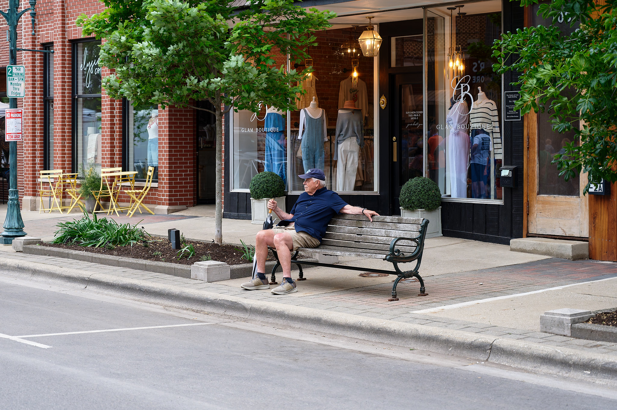 An elderly man sits on a bench in front of some storefronts while watching bike racing.