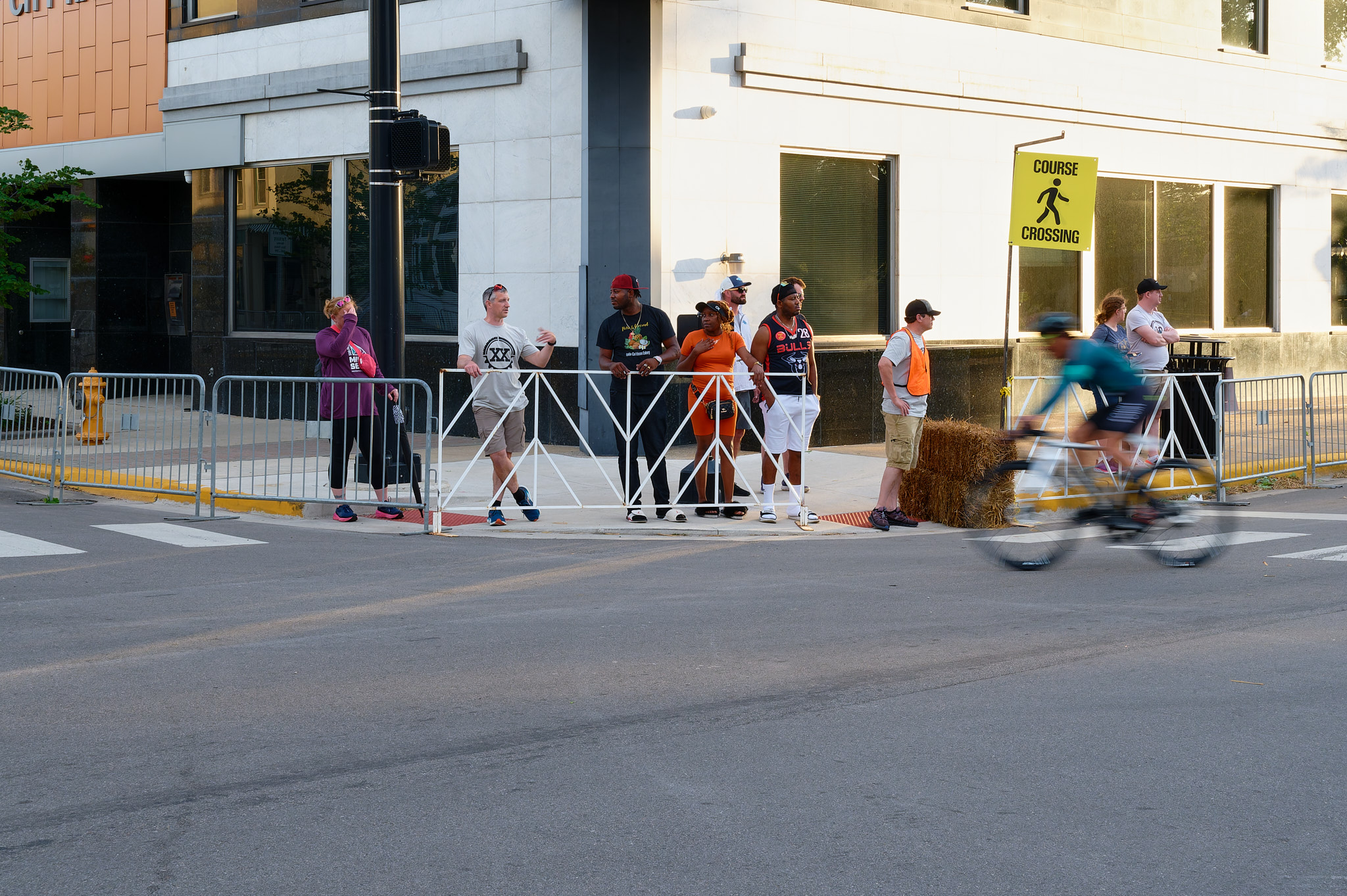 A bike racer speeds past a small crowd of spectators watching from the sidewalk.